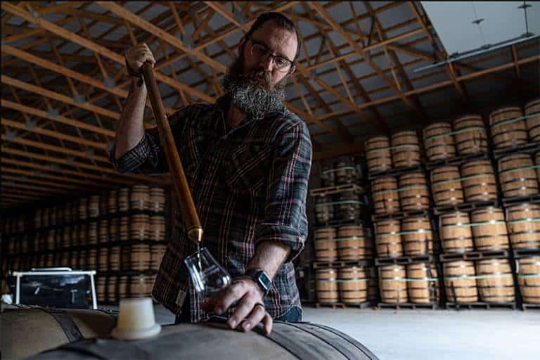 Distiller drawing whiskey from a barrel with a thief in a rickhouse